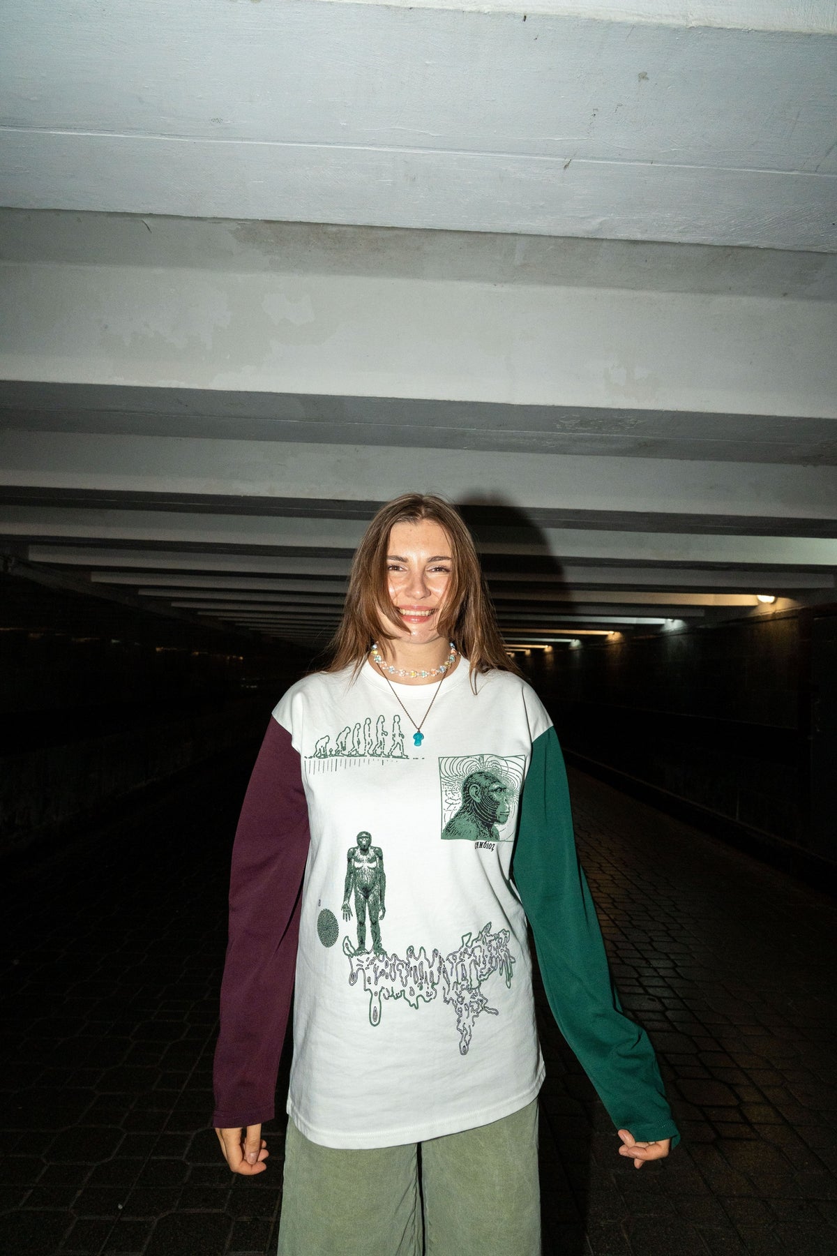 A young woman stands in a dark tunnel wearing a long-sleeve graphic shirt with a white body, a dark purple sleeve, and a dark green sleeve, paired with olive green shorts.