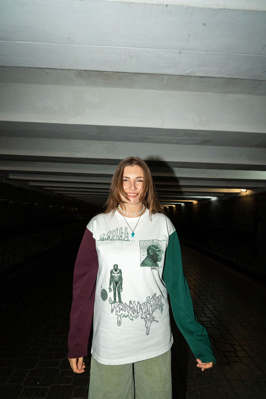 A young woman stands in a dark tunnel wearing a long-sleeve graphic shirt with a white body, a dark purple sleeve, and a dark green sleeve, paired with olive green shorts.