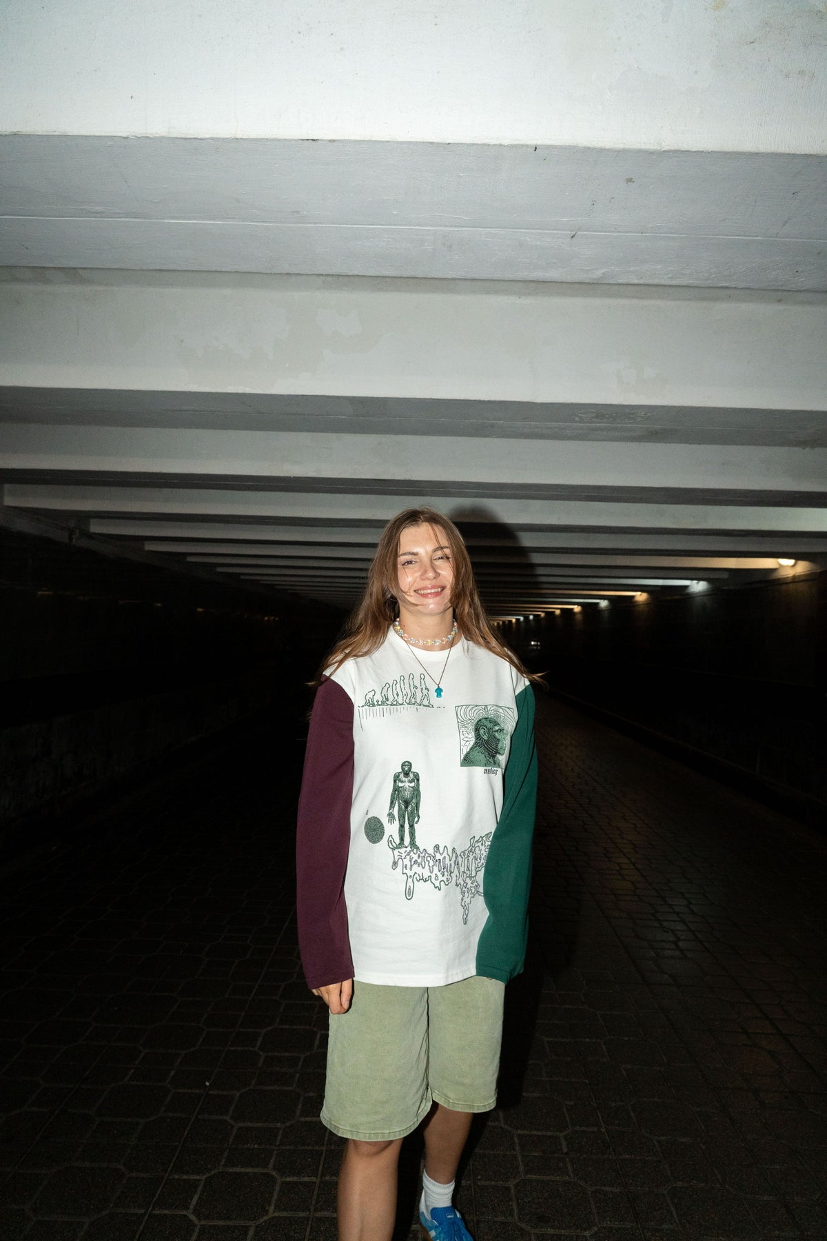 A woman standing in a dark tunnel wearing a white long-sleeve graphic shirt with maroon and green sleeves, olive green shorts, and blue sneakers.